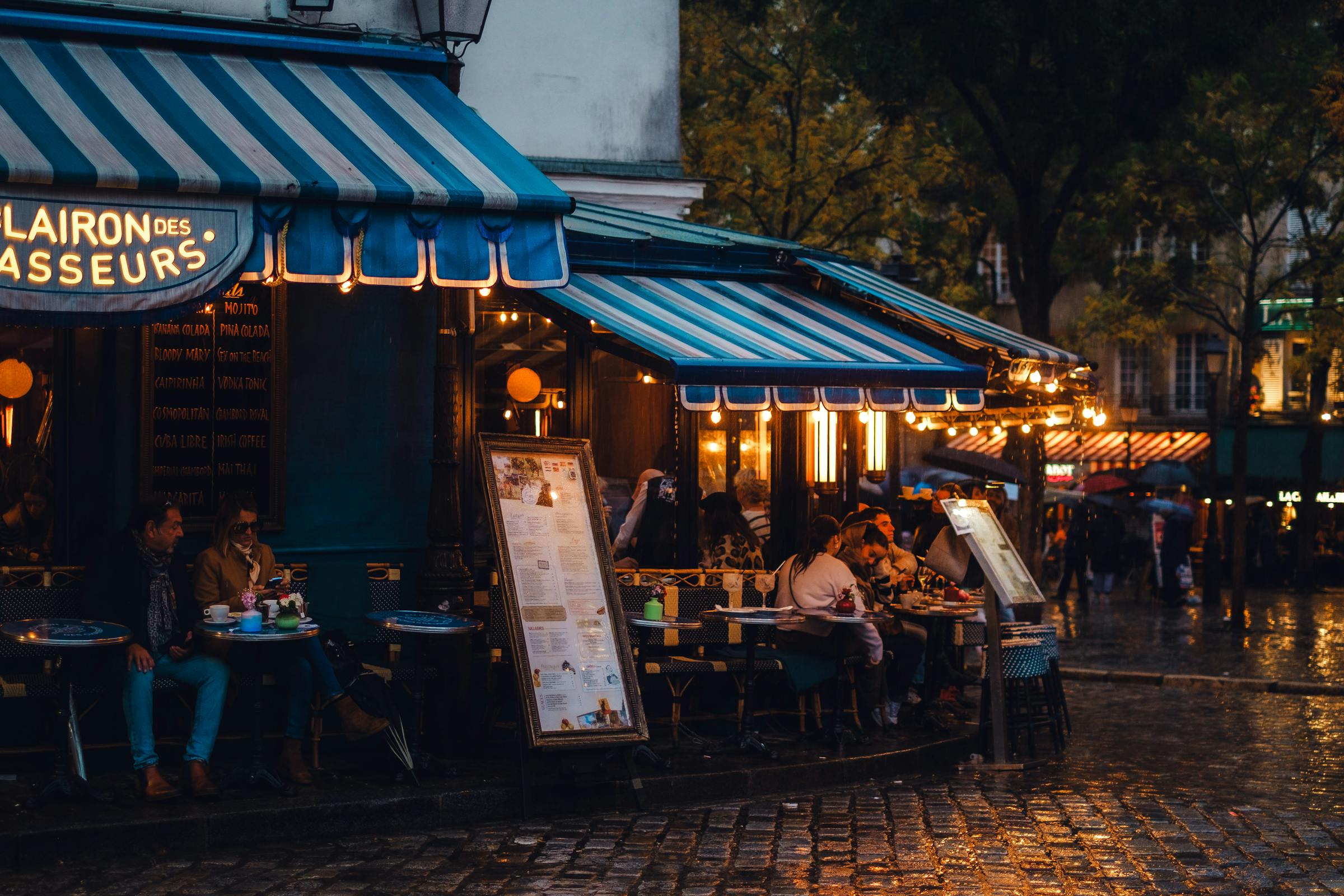 Regennasses Pariser Straßencafé mit warm beleuchteter Markise und Gästen am Abend